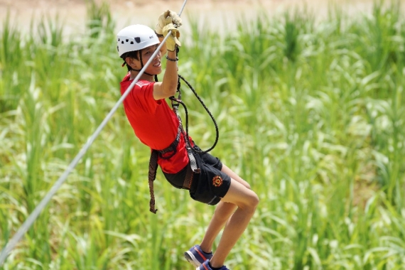 A wide shot of a male hanging in midair, wearing a red tee, black shorts, safety harness and a white helmet, ziplining his way across a maize field.