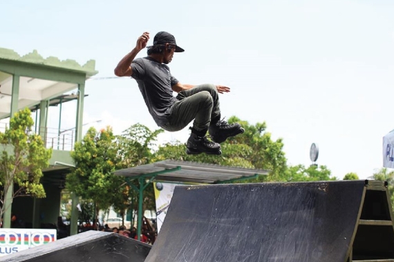 A wide shot of a male in action midair, dressed in charcoal grey ensemble, wearing a snapback and a pair of black rollerblades, cruising off a ramp.