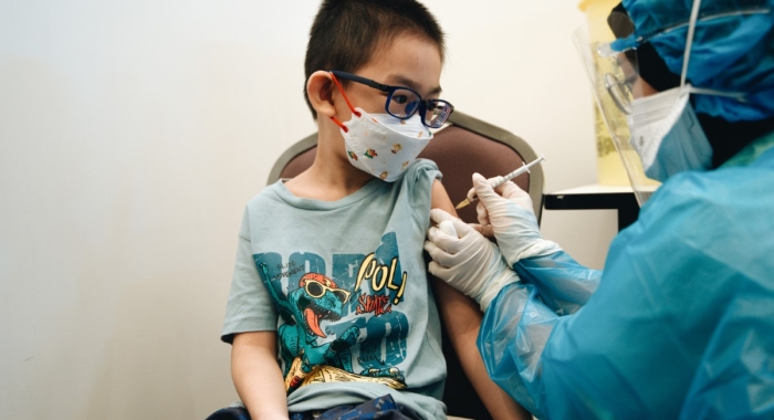 A medium landscape shot of a child in glasses boldly receiving his vaccination injection administered by a Sunway Medical Centre personnel dressed in complete protective gear