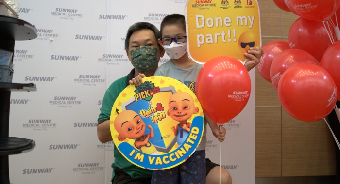 A medium full landscape shot of a boy posing for a commemoratory shot with his father, holding balloons and statement props after his first COVID-19 vaccination at Sunway Medical Centre