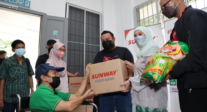 A medium full landscape shot of Datuk Patinggi Datuk Amar Puan Sri Jamilah Haji Anu as patron of Teratak Kasih Tok Nan (TKTN) alongside Sunway Group Brand Marketing and Communications CSR, Internal Branding & Events general manager Bernard Paul giving out essential groceries to beneficiaries at TKTN.