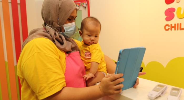 A tight medium landscape shot of a Sunway Little Sunshine nursery caregiver helping an infant check-in the Oodlins app upon arrival