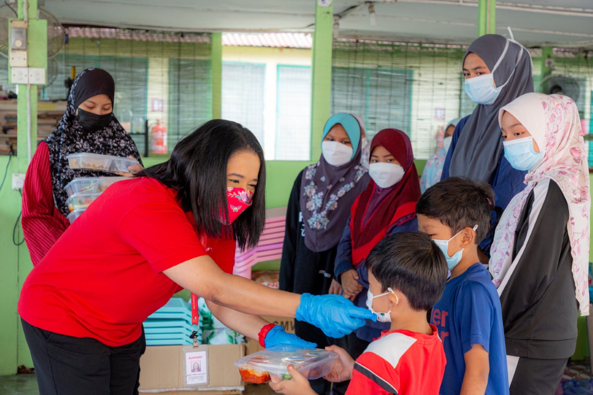 A lady in a red Sunway shirt giving packed meals to children wearing masks