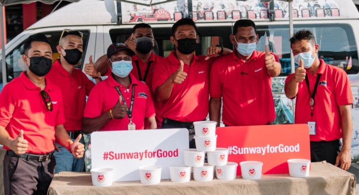 A medium landscape shot of Sunway volunteers in their classic red polo tee, flashing thumbs up behind #SunwayforGood signs propped up against bubur lambuk containers during Raya Cheer 2022.