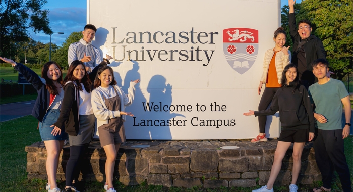 Evelyn Wee and some students posing in front of the Lancaster University signage, amidst greenery.
