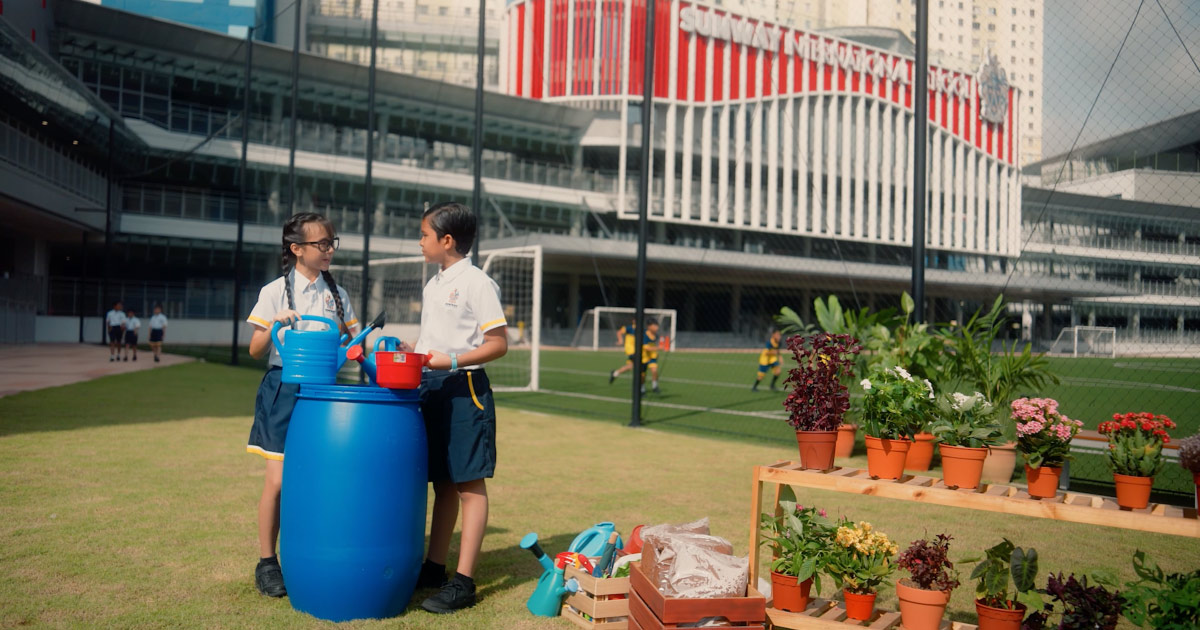 Two Sunway International School children at the Sunway International School football field, alongside plants and a water tank, with kids playing football in the background