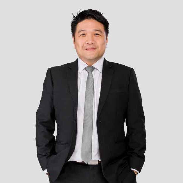 A portrait medium-shot of Sunway Group chief information officer Kevin Khoo Min Sinn, smirking at the camera with both hands tucked into his pants pocket, dressed in clasic black suit and a grey tie