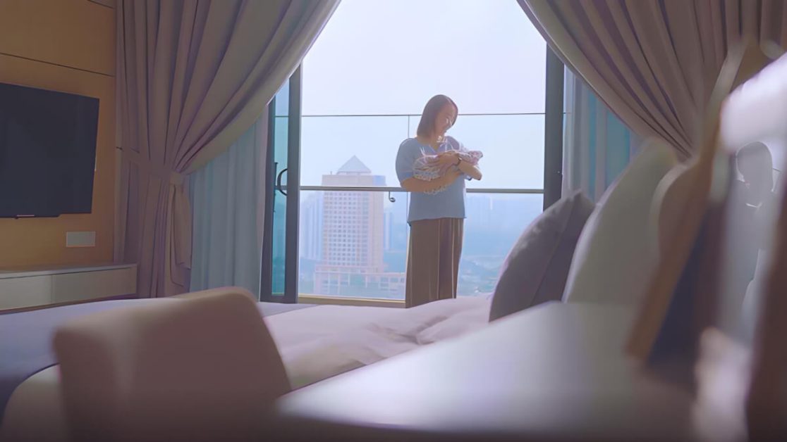 A mother and her baby standing by the window at the Postnatal Care Centre, facing Sunway Lagoon Hotel